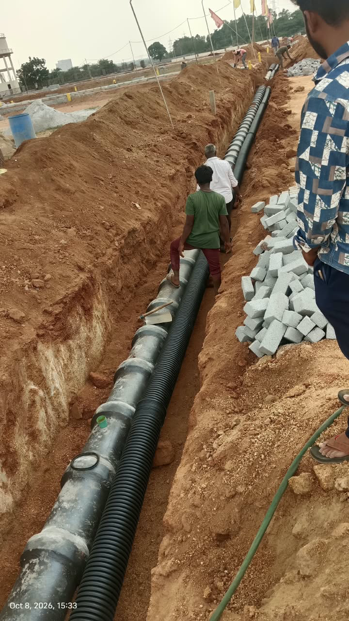 Twin underground pipelines — a ribbed double-wall pipe and a jointed pipe with branch ports — laid side by side in a deep road-side trench at Eastern Meadows, with workers jointing the line and kerb blocks stacked on the bank