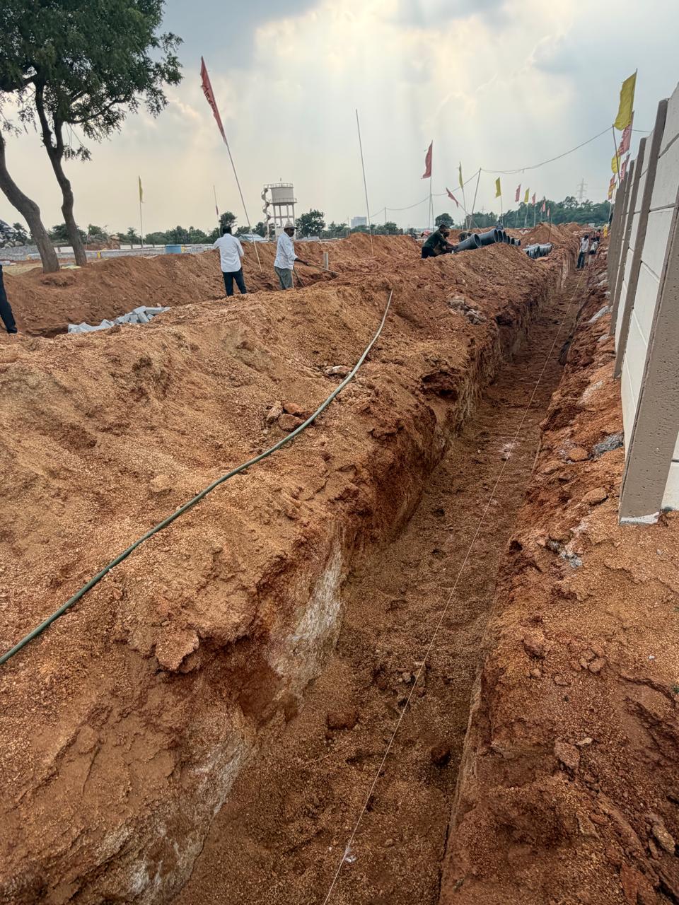 Deep pipeline trench cut along the compound wall at Eastern Meadows, set out with a string line, with the overhead water tank and a crew stacking pipes in the background