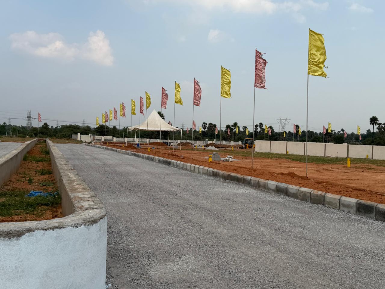 Kerb-lined road beside the planted median at Eastern Meadows, with levelled plots, yellow plot markers, the flag avenue and the compound wall on the right