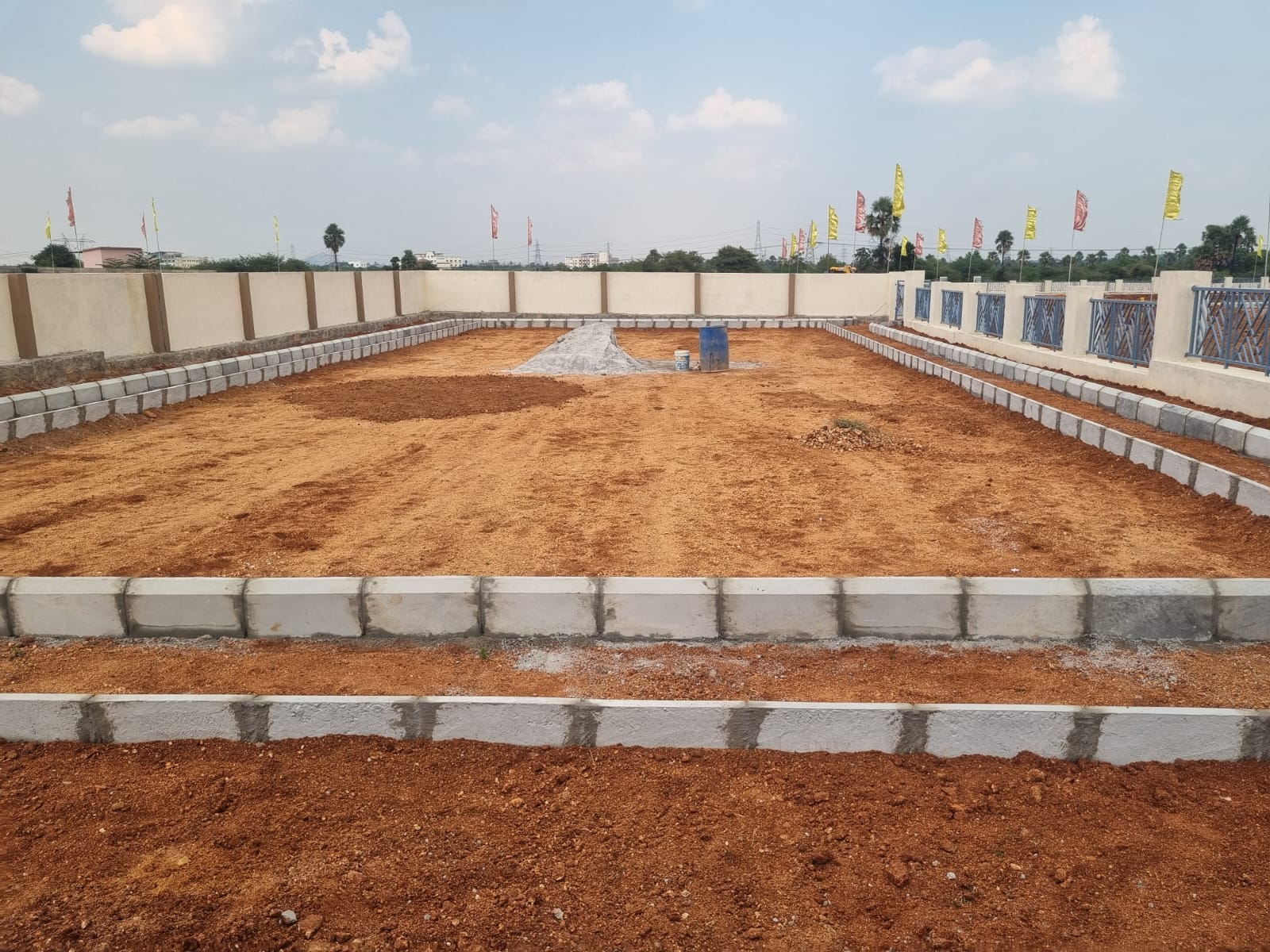 Front-on view of the jogging track under construction at Eastern Meadows: a kerb-edged loop on levelled red soil, between the precast compound wall and the decorative grille boundary wall