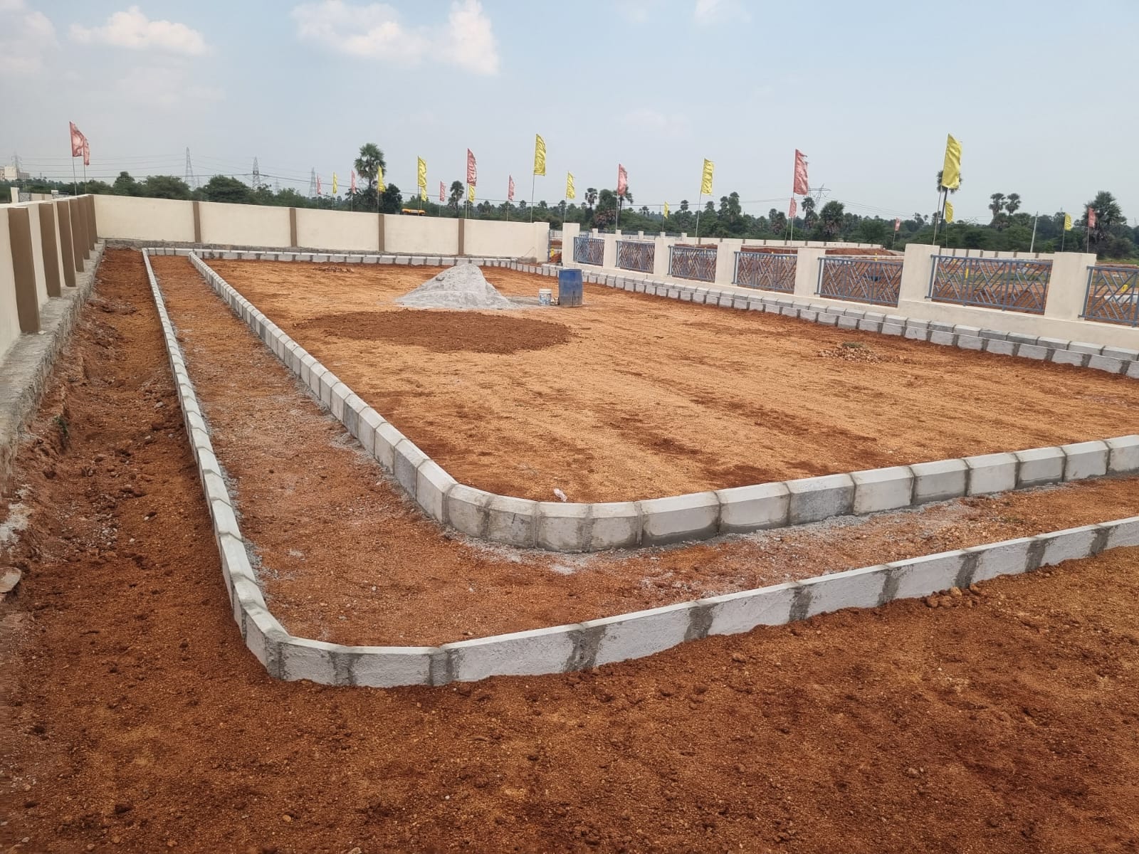 Jogging track under construction along the compound wall at Eastern Meadows, its path marked out by a double row of kerb stones on graded red soil, with a decorative grille wall alongside