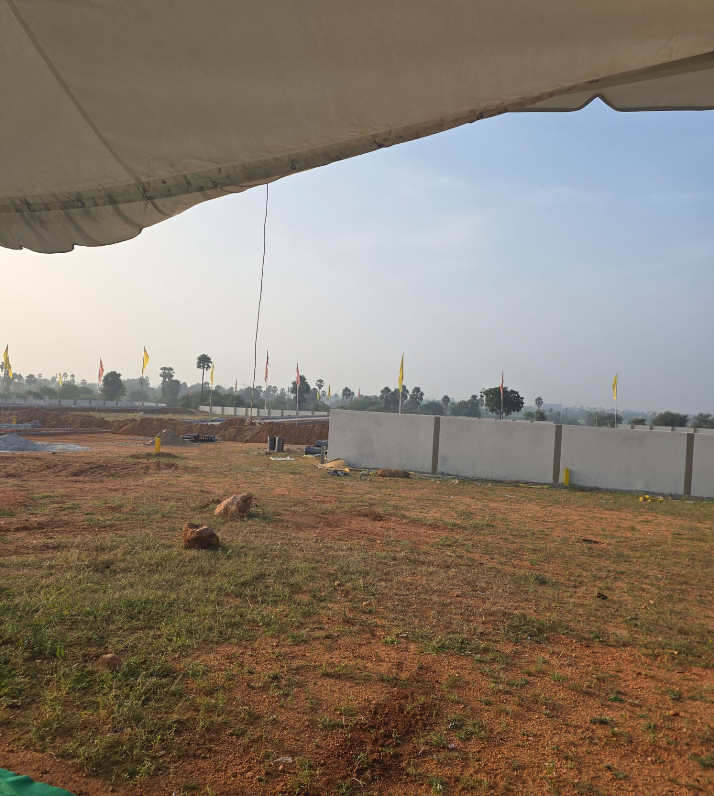 Open plotted land at Eastern Meadows seen from the site-visit tent, with a run of compound wall and the internal road beyond