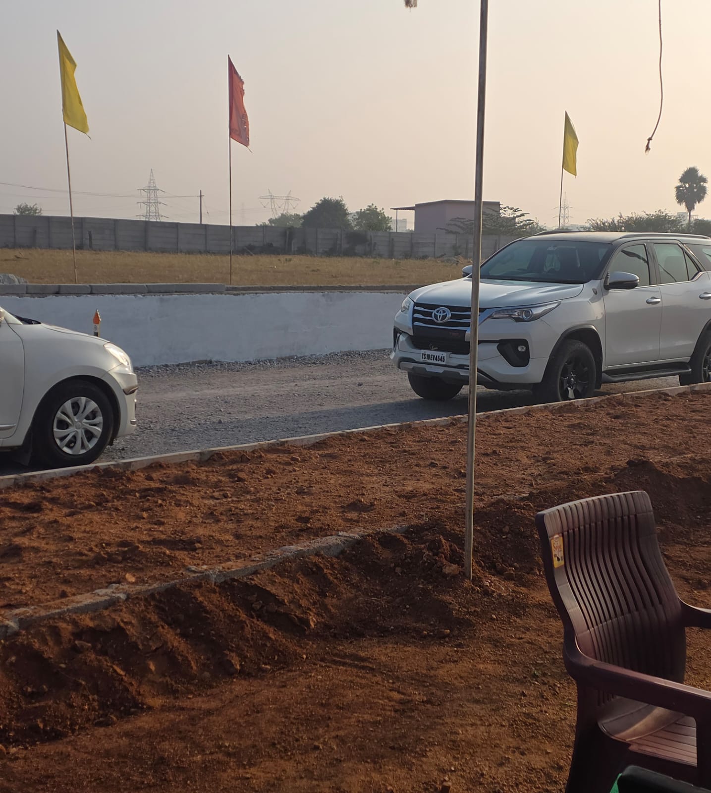 Entrance road at Eastern Meadows lined with flags, visitor cars parked, and a freshly dug landscaping bed in the foreground